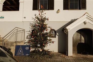 Ein mit Ornamenten geschmückter Weihnachtsbaum steht vor einem Gebäude. Das Gebäude hat weiße Wände, eine Holztür und eine Treppe. Ein blaues Schild mit weißen Schriftzeichen befindet sich neben dem Baum.