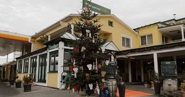 Ein kleiner Weihnachtsbaum steht vor einem gelben Gebäude mit dem Schild 'Café & Restaurant'. Um den Baum herum stehen Topfpflanzen und Fässer. Der Himmel ist bedeckt.