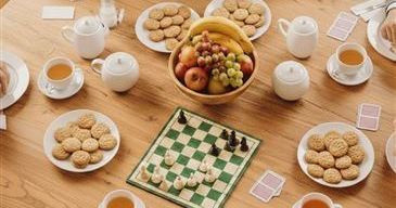 Ein Holztisch ist mit Tellern voller Kekse, einer Obstschale, einem Schachspiel und Tassen Tee gedeckt. Der Tisch ist von Menschen umgeben, die eine Mahlzeit genießen und Schach spielen.