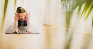 Eine Frau in einem rosa Oberteil und blauen Leggings praktiziert Yoga auf einer Matte, beugt sich mit den Händen auf dem Boden nach vorne in einer häuslichen Umgebung.