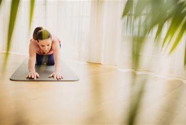 Eine Frau in einem rosa Oberteil und blauen Leggings praktiziert Yoga auf einer Matte, beugt sich mit den Händen auf dem Boden nach vorne in einer häuslichen Umgebung.