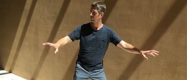 Ein Mann in einem blauen T-Shirt und Jeans steht mit ausgestreckten Armen in einem Raum und wirft einen Schatten auf einen hellen Boden und eine beige Wand.