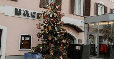 Ein geschmückter Weihnachtsbaum steht vor einem rosa Gebäude mit der Aufschrift 'BACH'. Ein blauer Schild mit '21' steht neben dem Baum. Das Gebäude hat mehrere Fenster und einen Glaseingang. Ein roter Stuhl ist im Eingangsbereich zu sehen.