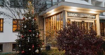 Ein mit Ornamenten geschmückter Weihnachtsbaum steht vor dem Simon des Vitalotel, einem Hotel mit Glasfenstern und einem gut beleuchteten Eingang.