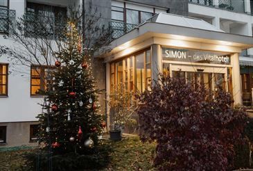 Ein mit Ornamenten geschmückter Weihnachtsbaum steht vor dem Simon des Vitalotel, einem Hotel mit Glasfenstern und einem gut beleuchteten Eingang.
