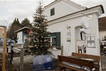 Ein weißes Haus mit einem Weihnachtsbaum davor, geschmückt mit bunten Ornamenten und einem blauen Schild mit der Nummer 17. In der Nähe sind Holzbänke und ein Holzzaun. Ein Auto ist auf der Straße geparkt.