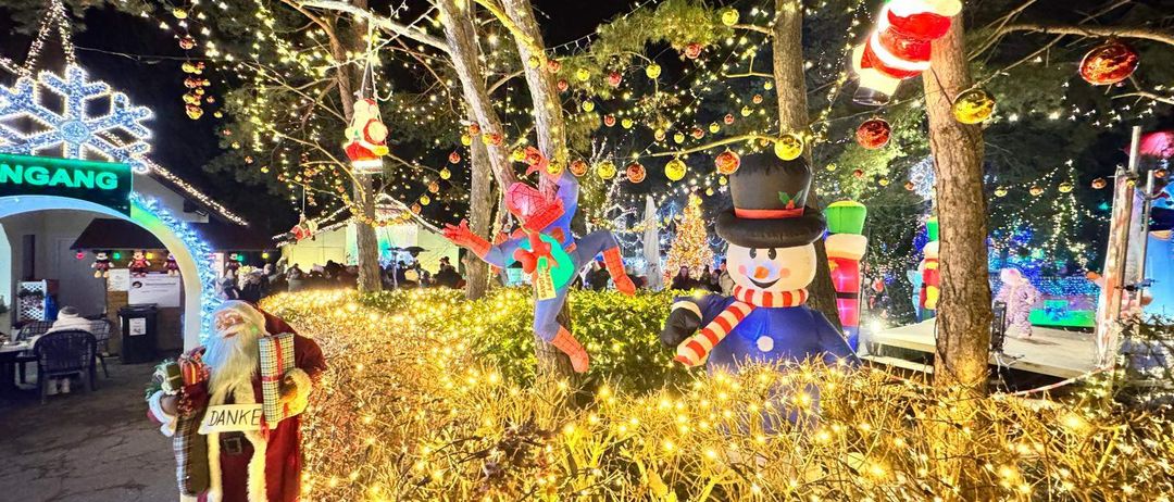 Ein nachtlicher Weihnachtspark mit Spiderman- und Weihnachtsmannfiguren, beleuchteten Baumen und bunten Lichtern.