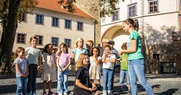 Eine Gruppe von Kindern und zwei Frauen stehen vor einem Gebäude mit vielen Fenstern. Die beiden Frauen unterrichten die Kinder. Die Frau auf der rechten Seite hält einen Stock.