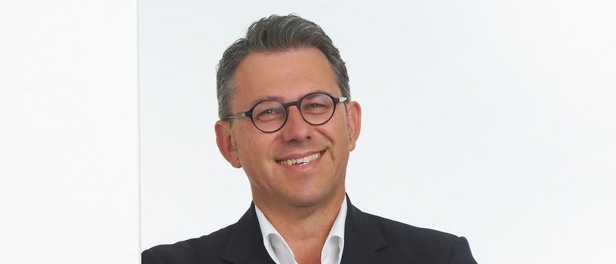 Ein Mann in einem schwarzen Anzug und mit Brille lächelt und posiert mit verschränkten Armen vor einer weißen Wand.