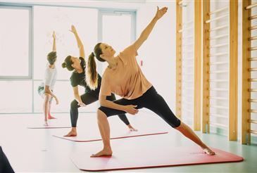 Vier Frauen praktizieren Yoga-Posen in einem Raum mit Holzwänden, Matten und einem Glasfenster. Eine Frau hebt die Arme, eine andere beugt sich nach vorne und zwei andere strecken die Beine. Der Raum ist hell.