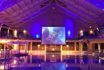 Ein Hallenbad mit einer großen Projektionsleinwand, die eine Karte anzeigt. Der Bereich ist mit lila Lichtern beleuchtet, mit Liegestühlen und Tischen in der Nähe.