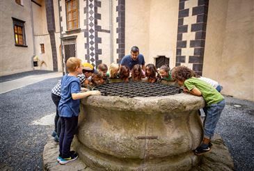 Kinder versammeln sich um einen großen steinernen Brunnen, blicken durch ein Gitter, während ein erwachsener Mann in einem Innenhof steht.