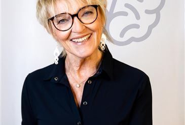 Eine lächelnde Frau mit kurzen blonden Haaren, die eine Brille und ein schwarzes Hemd trägt, posiert für ein Foto mit weißem Hintergrund.