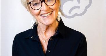 Eine Frau mit kurzen blonden Haaren lächelt und trägt eine Brille, ein schwarzes Hemd und eine Halskette. Sie steht vor einer weißen Wand mit einer grauen gehirnähnlichen Illustration.