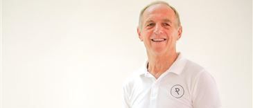 Ein älterer Mann in einem weißen Poloshirt mit einem Logo auf der linken Brustseite lächelt und scheint zu tanzen. Er hat einen fröhlichen Ausdruck und befindet sich wahrscheinlich in einem Tanzstudio.