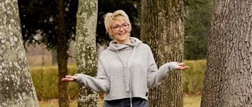 Eine Frau mit kurzen blonden Haaren und Brille steht mit ausgestreckten Armen zwischen zwei großen Bäumen. Sie trägt eine graue Kapuzenjacke und weite Hosen. Der Hintergrund zeigt ein Feld mit weiteren Bäumen.