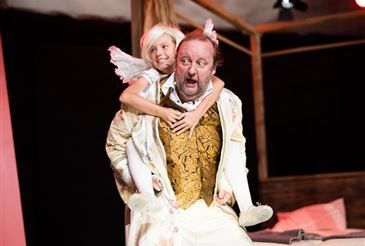 Ein Mann in aufwendiger Kleidung hält ein kleines Mädchen auf seinen Schultern. Sie scheinen auf einer Bühne zu sein. Das Mädchen trägt Engelsflügel und ein rosa Kleid. Der Mund des Mannes steht offen.