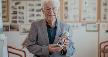 Ein älterer Mann in einem grauen Anzug steht in einem Raum und hält ein buntes Buch in der Hand, mit gerahmten Bildern und Stühlen im Hintergrund.