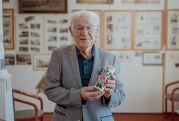 Ein älterer Mann mit weißem Haar und Brille steht in einem Raum und hält ein kleines Objekt in der Hand. Hinter ihm befinden sich mehrere gerahmte Bilder an der Wand und Stühle im Hintergrund.