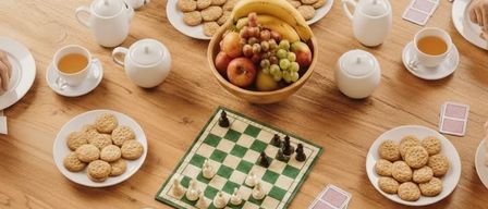 Eine Gruppe von Menschen sitzt um einen Holztisch herum mit einem Schachbrett, Keksen und einer Schüssel Obst. Auf dem Tisch stehen Tassen, Teekannen und Untertassen.