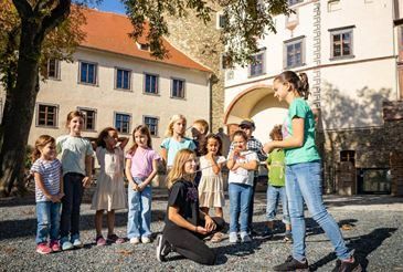 Eine Gruppe von Kindern und zwei Frauen steht vor einem Gebäude mit Bogenfenstern, eine Frau kniet und zeigt den Kindern etwas.