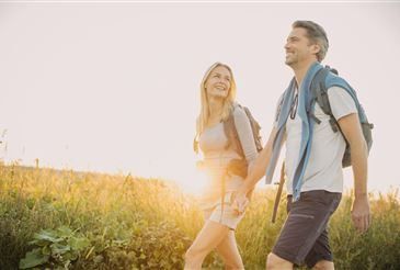 Ein Mann und eine Frau gehen Hand in Hand durch ein saftiges Feld. Der Mann trägt einen Rucksack und die Frau trägt ein Kleid. Sie lächeln in Richtung untergehender Sonne.