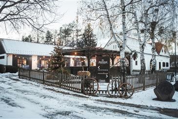 Ein rustikales Haus mit schneebedecktem Dach, umgeben von einem Holzzaun und Bäumen, mit einem Weihnachtsbaum und dekorativen Rädern davor.