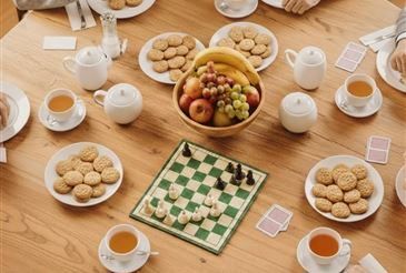 Ein Holztisch ist mit verschiedenen Lebensmitteln, Getränken und einem Schachbrett bestückt. Teller mit Keksen, eine Obstschale und Teekannen umgeben das Schachbrett. Mehrere Tassen Tee stehen auf dem Tisch, und Spielkarten sind verstreut.