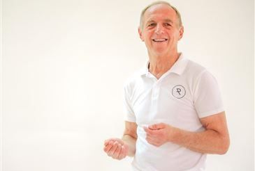 Ein älterer Mann in einem weißen Poloshirt mit Logo lächelt, während er in einem weißen Raum steht. Er hat die Hände erhoben und scheint zu tanzen.