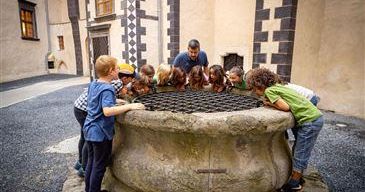 Eine Gruppe von Kindern und einem Erwachsenen schaut in einen großen steinernen Brunnen auf einem Freigelände.