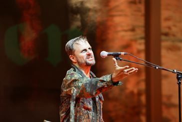 Ein Mann in einer Camouflage-Jacke steht auf der Bühne, singt in ein Mikrofon und hat ein verschwommenes Publikum im Hintergrund.