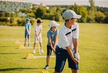 Fünf Kinder spielen an einem sonnigen Tag Golf. Sie sind in Golfbekleidung gekleidet und halten Golfschläger. Es gibt mehrere gelbe und weiße Golfbälle auf dem Gras.
