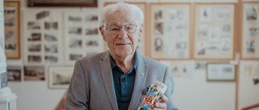 Ein älterer Mann mit Brille steht in einem Raum und hält eine bunte Karte in der Hand. Er ist in einem grauen Anzug gekleidet. Hinter ihm hängen mehrere gerahmte Bilder an einer weißen Wand.