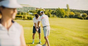 Zwei Golfer befinden sich auf einem sonnigen Golfplatz, einer gibt dem anderen eine Lektion, während eine Frau von hinten zusieht.