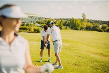 Zwei Golfer befinden sich auf einem sonnigen Golfplatz, einer gibt dem anderen eine Lektion, während eine Frau von hinten zusieht.