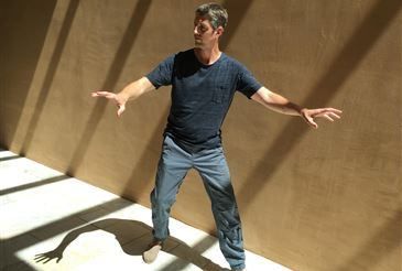 Ein Mann in einem blauen T-Shirt und Jeans steht mit ausgestreckten Armen in einem Raum mit einer beigen Wand.