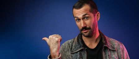 Ein Mann mit Bart und Schnurrbart in einem schwarzen T-Shirt und einer Jeansjacke posiert vor einem blauen Hintergrund und zeigt mit dem Zeigefinger nach oben.