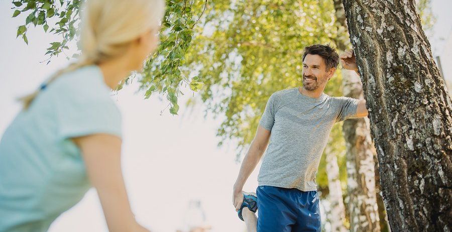 Ein Mann und eine Frau stehen in einem Park. Der Mann lehnt sich gegen einen Baum, lächelt und hält eine Wasserflasche. Die Frau steht ihm gegenüber, hält eine Wasserflasche und schaut ihn an.