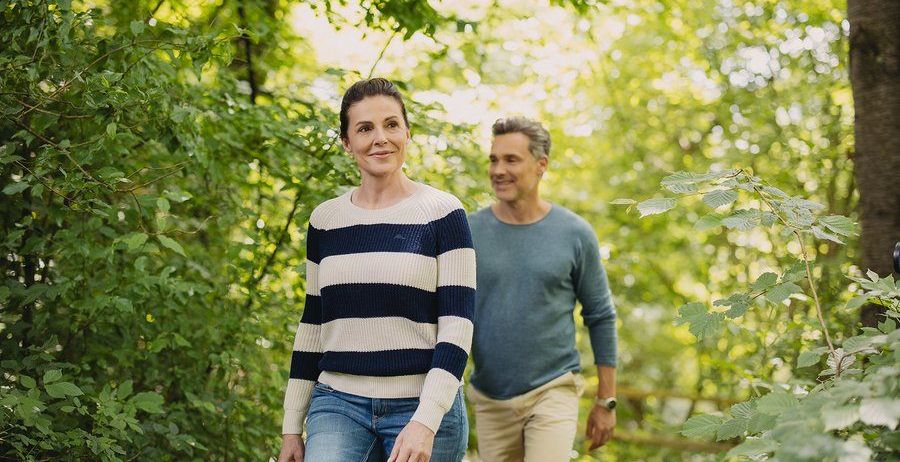 Eine lächelnde Frau und ein Mann gehen gemeinsam in einem üppigen, grünen Wald. Die Frau trägt einen gestreiften Pullover und Jeansshorts, während der Mann ein blaues Hemd und Khaki-Shorts trägt.