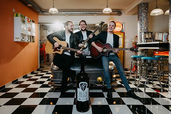 Drei Männer in einem Raum mit einem karierten Boden, jeder hält eine Gitarre, posieren mit einer Flasche Blue Franks vor einem Oldtimer.