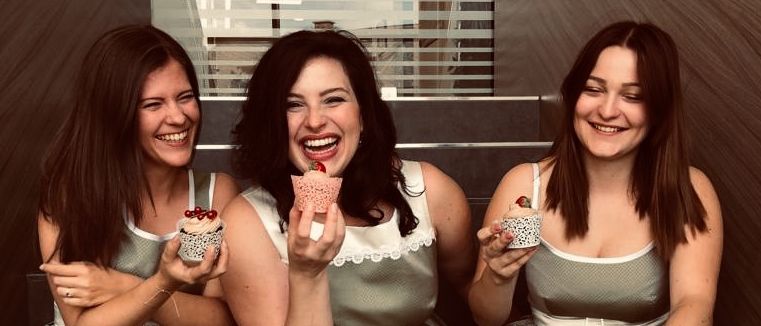 Drei Frauen in passenden Kleidern, die Cupcakes halten, zusammen sitzen und in die Kamera lächeln.