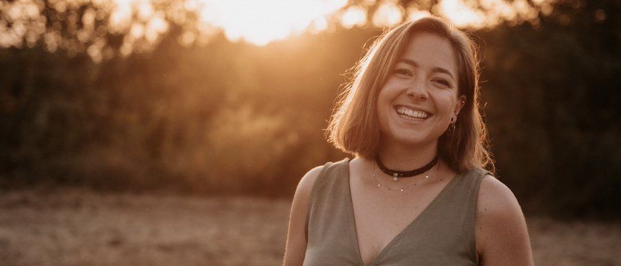 Eine Frau mit kurzen Haaren lächelt auf einem Feld mit einem Sonnenuntergang im Hintergrund. Sie trägt ein ärmelloses Kleid und hat eine Tätowierung am Arm.
