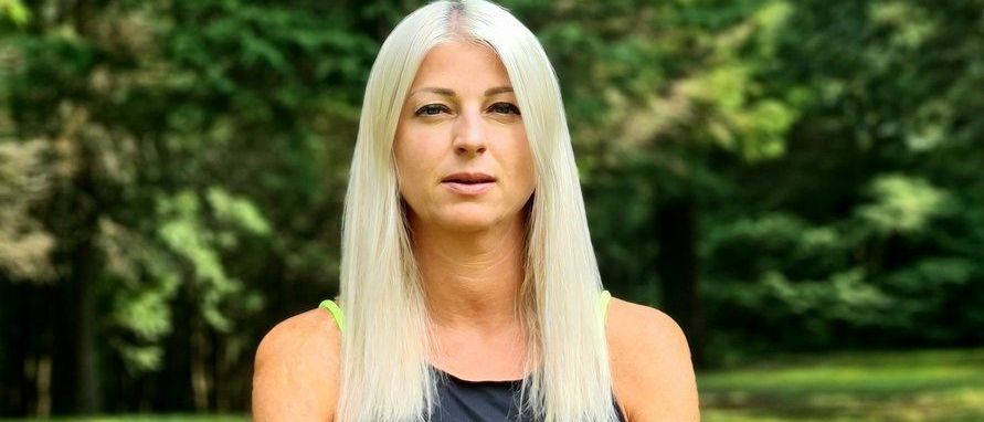 Eine Frau mit langen blonden Haaren sitzt auf dem Gras und praktiziert Yoga in einem Park. Sie trägt ein schwarzes Tanktop und hat ihre Hände in einer Meditationshaltung.