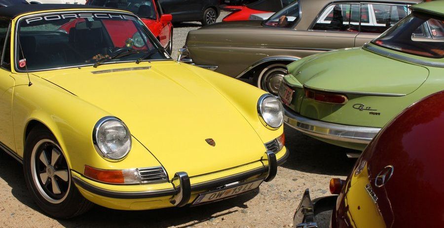 Ein gelber Porsche 911 Sportwagen ist unter anderen Oldtimern auf einem Kiesplatz geparkt.