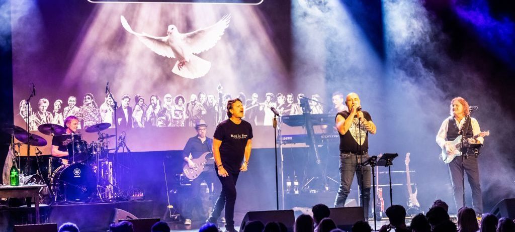 Ein Live-Konzert mit einer Band auf der Bühne, eine Taube fliegt oben, und eine große Leinwand hinter ihnen mit dem Text 'Rock in Peace'. Die Bandmitglieder spielen vor einem Publikum.
