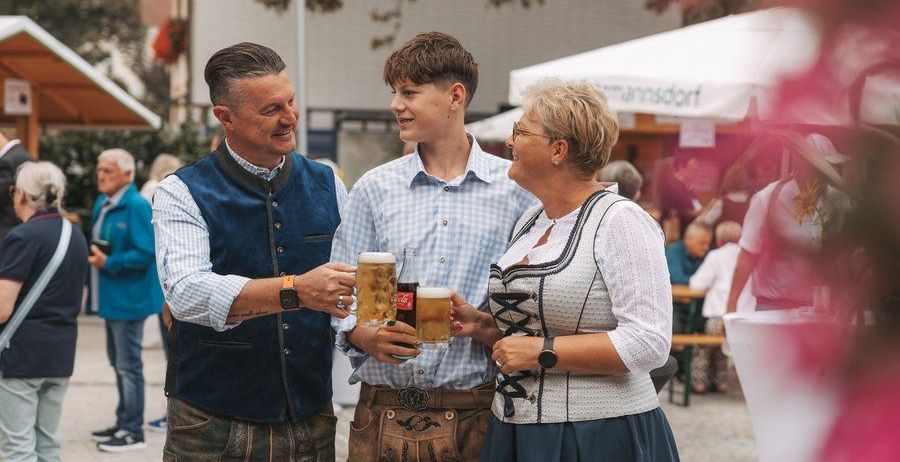 Drei Personen in traditioneller deutscher Kleidung stehen zusammen und halten Gläser mit Bier in der Hand. Der Mann links trägt eine blaue Weste, der Mann in der Mitte trägt ein kariertes Hemd und die Frau rechts trägt ein weißes Oberteil mit einem blauen Rock. Sie lächeln und scheinen sich auf einem Festival zu amüsieren.