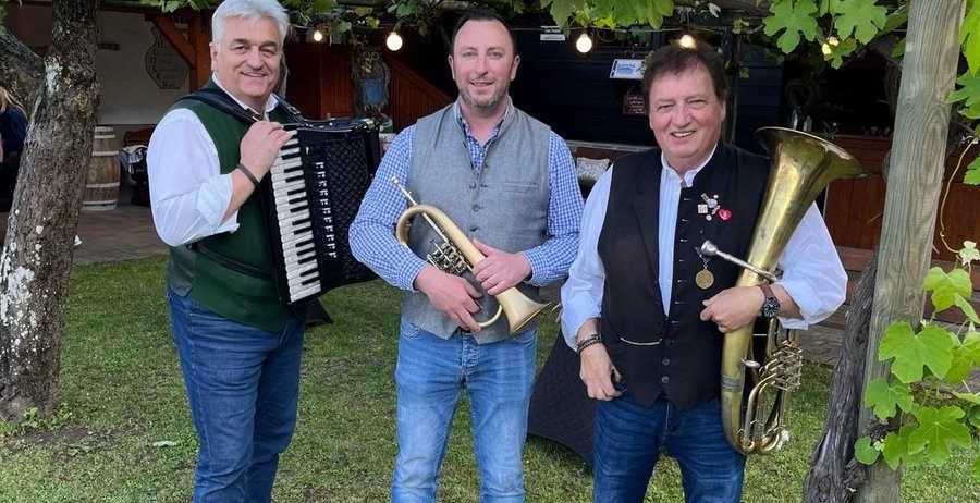 Drei Männer stehen in einem Garten, jeder hält ein Musikinstrument. Der Mann auf der linken Seite spielt Akkordeon, der in der Mitte eine Trompete und der auf der rechten Seite eine Tuba. Sie alle lächeln.