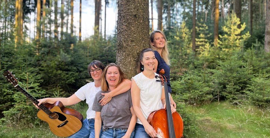 Vier Musiker posieren für ein Foto im Wald, einer hält eine Gitarre und ein anderer ein Cello. Sie lächeln und lehnen sich gegen einen Baumstamm.