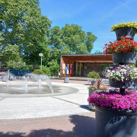 Ein öffentlicher Park mit einem Brunnen, Bäumen und einem kleinen Holzgebäude. Bunte Blumen in großen Töpfen rund um den Brunnen und das Gebiet ist von gepflasterten Ziegeln umgeben.
