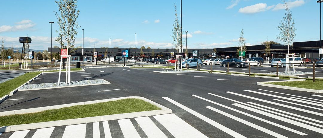 Ein leerer Parkplatz mit weißen Markierungen und Grasflächen. In der Ferne befinden sich Gebäude, Autos und Bäume. Der Himmel ist blau mit einigen Wolken.